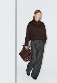 Donna che indossa una giacca bottonata marrone scuro, pantaloni a plettro grigio a righe larghe, scarpe nere, e porta una grande borsa a tracolla marrone scuro con texture.
