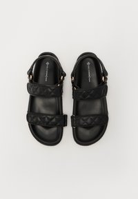 Sandales véganes noires avec deux brides matelassées ornées d'accents en métal doré. Le design présente une semelle plate et un style à dos ouvert.