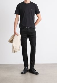 Czarna bawełniana koszulka z małym logo, w zestawie z czarnymi dżinsami i czarnymi skórzanymi butami. Trzyma jasnobeżową kurtkę dżinsową.