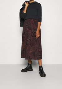 Sweat à capuche noir avec cordon, associé à une longue jupe marron ornée d'un motif rouge ondulé. Bottines noires avec une semelle épaisse et des détails de surpiqûres.