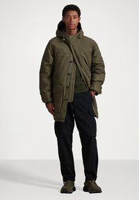 Casaco acolchoado verde oliva com capuz, grandes bolsos frontais e fecho de correr, combinado com uma camisola de malha verde escura e calças cargo pretas.