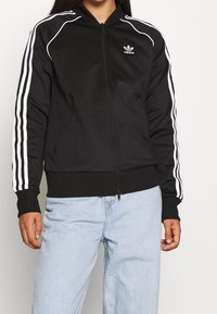 Černá sportovní bunda na zip s bílým designem tří pruhů na rukávech, vybavená žebrovaným límcem a manžetami a bočními kapsami.