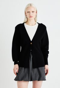 Black cardigan in a soft knit, featuring a deep V-neck, single button closure, and long, slightly puffed sleeves. Paired with a striped skirt.