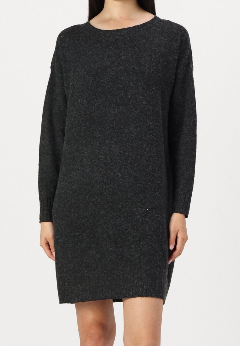 Femme portant une robe pull en maille gris foncé à manches longues avec un col rond sur un fond clair uni.