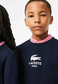 Sweat-shirt bleu marine avec un logo Lacoste blanc et le texte "PARIS". Le col est rose. Les cheveux du modèle sont tressés et coiffés sur le côté.