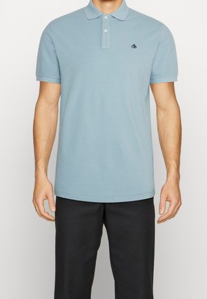 Un homme porte un polo à manches courtes bleu clair avec un petit logo sombre sur la poitrine, assorti à un pantalon noir, sur un fond uni.