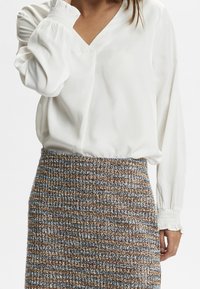 Blouse blanche à col en V et manches bouffantes, associée à une jupe mi-longue texturée avec un motif tissé multicolore.