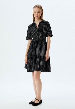 Vestido negro con lunares, con un cuello tipo camisa a botones, mangas cortas, cintura fruncida y falda con volantes. Se lleva con zapatos planos negros.