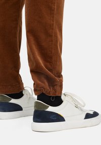 Witte en marineblauwe sneakers met groene accenten, textuurmaterialen en veters. Bruine corduroy broek met ribbelstructuur bij de zoom.