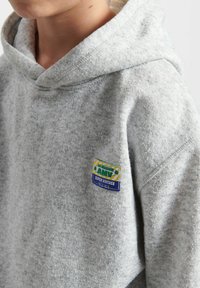 Felpa grigia con cappuccio realizzata in un tessuto testurizzato. Presenta un'etichetta colorata sul petto con testo ed elementi grafici.