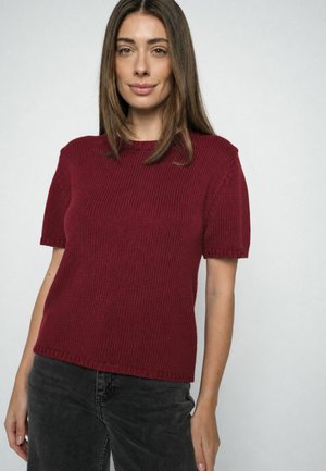 Žena s rovnými hnedými vlasmi, oblečená v krátkosrstý burgundský pletený sveter a tmavomodré džínsy na jednoduchom svetlom pozadí.
