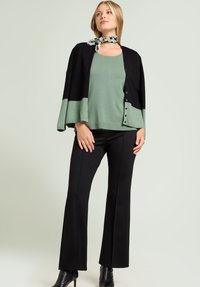 Vrouw die een zwarte en saliegroene cardigan draagt over een saliegroen topje, zwarte broek, zwarte laarzen met hak en een patroonige nek sjaal, zelfverzekerd staand.