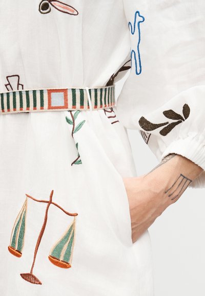 Άτομο που φοράει λευκό ένδυμα με κεντημένα σε πράσινο και πορτοκαλί σύμβολα κλίμακας, ριγωτή ζώνη και χέρι με τατουάζ γραμμές στην τσέπη.