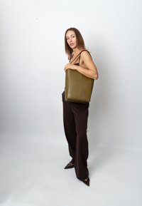 Olive green croc-embossed tote bag with a rectangular shape. Solid material and textured surface. Worn on shoulder; brown wide-legged pants.
