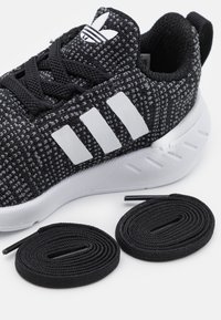 Sneaker lavorato a maglia in nero e grigio con marchio a tre strisce bianche, suola bianca arrotondata e due lacci neri. Parte superiore testurizzata, design atletico.