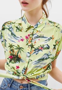 Kortärmad, knappad skjorta i ljusgrön tyg, med ett tropiskt mönster av blommor, berg och palmträd, knuten vid midjan.