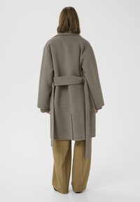 Abrigo de lana gris con un cinturón en la cintura, mangas anchas y aberturas laterales, combinado con pantalones sueltos color beige. Diseño limpio y minimalista.