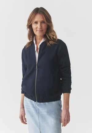 Marineblauer Bomberjacke mit Reißverschluss, gerippten Bündchen und Saum. Getragen über einem weißen Hemd mit Kragen und kombiniert mit einem hellen Jeansrock.