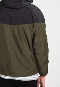 Olive green jacket with a black upper section and hood, featuring a smooth texture, elastic cuffs, and a straight hem at the waist.