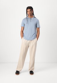 Camiseta de manga corta azul claro combinada con pantalones beiges de corte suelto y zapatillas negras con suelas marrones, contra un fondo blanco.