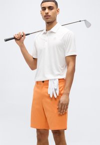 Άνδρας με λευκό πόλο και πορτοκαλί σορτς κρατώντας ένα μπαστούνι του γκολφ στον ώμο με λευκά γάντια κρυμμένα στην τσέπη, μπροστά σε απλό φόντο.