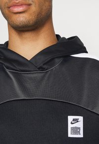 Felpa nera con cappuccio, caratterizzata da una texture di tessuto liscio, e un'etichetta logo bianca con scritto "NIKE FORCE" sul davanti.