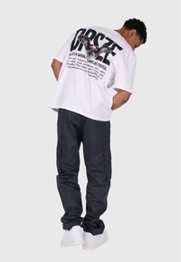 Hvit oversized t-skjorte med grafisk tekst og fugledesign på baksiden, kombinert med sorte cargo-bukser og hvite sneakers.