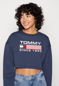 Sudadera recortada azul marino de manga larga, con un logo blanco de "TOMMY" y un diseño de bandera bordado. Combinada con jeans de mezclilla azul claro.