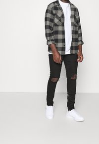 Person i en svart-grå rutigt skjorta, vit t-shirt, slitna svarta skinny jeans och vita sneakers som står mot en enfärgad bakgrund.