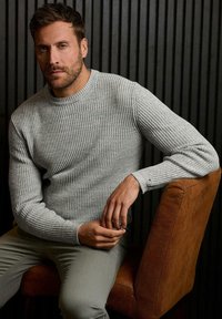 Helles graues geripptes Sweatshirt mit einem Rundhalsausschnitt, strukturiertem Gewebe und langen Ärmeln, getragen über hellgrauen Hosen, während auf einem braunen Stuhl gesessen wird.