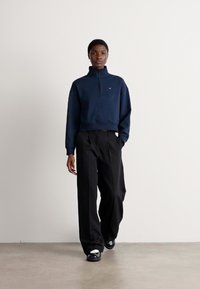 Navy zip-neck sweatshirt with ribbed cuffs, paired with black wide-leg trousers and black slip-on shoes against a neutral background.