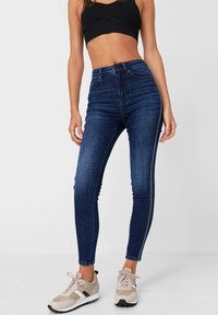 Los jeans ajustados de color azul oscuro presentan una cintura alta, una textura suave y un detalle de cremallera en contraste a lo largo del costado. Se llevan con zapatillas en tonos neutros.