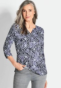 Langarmshirt mit V-Ausschnitt, das ein geometrisches Muster in Blau- und Lavendeltönen zeigt, aus glattem Stoff gefertigt. Graue Jeans getragen.