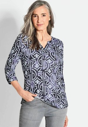 Langarmshirt mit V-Ausschnitt, das ein geometrisches Muster in Blau- und Lavendeltönen zeigt, aus glattem Stoff gefertigt. Graue Jeans getragen.