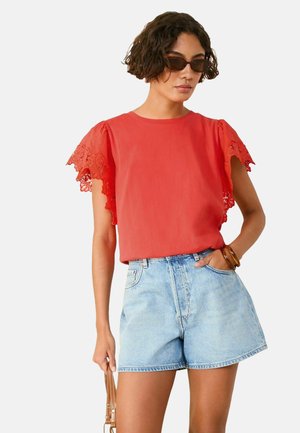 Maglietta rossa con maniche in pizzo, abbinata a pantaloncini in denim azzurro chiaro. La maglietta è a tinta unita con scollatura rotonda, mentre i pantaloncini hanno una vestibilità comoda.