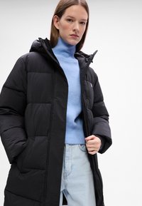 Piumino nero con cappuccio, caratterizzato da un design trapuntato e tasche laterali, indossato sopra un maglione azzurro chiaro e jeans in denim.