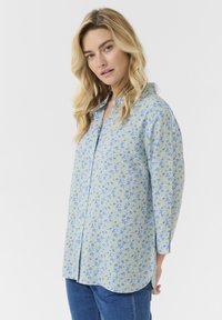Camicia azzurra chiara con un motivo floreale giallo e blu, maniche lunghe e orlo curvo. Realizzata in tessuto morbido.