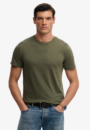 Homme portant un t-shirt à manches courtes vert olive, jean bleu, ceinture noire et montre-bracelet avec un bracelet en cuir marron, mains dans les poches.
