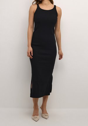 Femme portant une robe midi noire sans manches avec des fentes latérales et des chaussures beige pointues à enfiler, debout devant un fond uni.