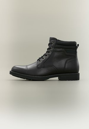Bottine noire en cuir avec laçage à l'avant, col matelassé, bout rond et petit talon sur fond neutre.