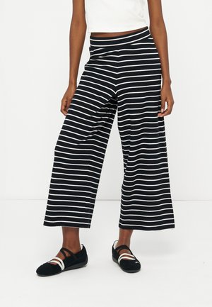 Γυναίκα με μαύρο παντελόνι φαρδύ, κομμένο στο ύψος του αστραγάλου, με λευκές οριζόντιες ρίγες και μαύρα μπαλέτα με λευκές λουρίδες, σε λευκό φόντο.