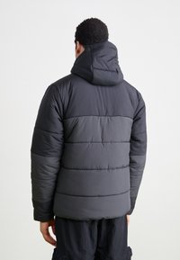 Vadderad svart jacka med huva, med horisontell sömnad och texturerat material. Baksidan visar en strömlinjeformad siluett och mjuk finish.