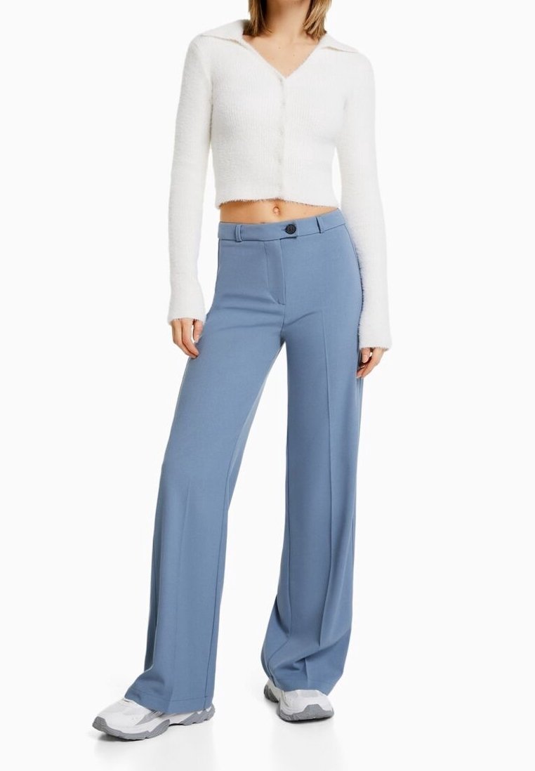 Femme portant un haut blanc court à manches longues boutonné, un pantalon large bleu clair taille haute et des baskets blanches, debout les mains détendues.
