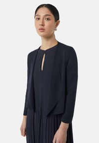 Marineblauer Cardigan, leichtes Strickgewebe, offene Vorderseite mit drapiertem Verschluss, lange Ärmel, kombiniert mit einem plissierten Kleid. Glatte Textur, minimales Design.