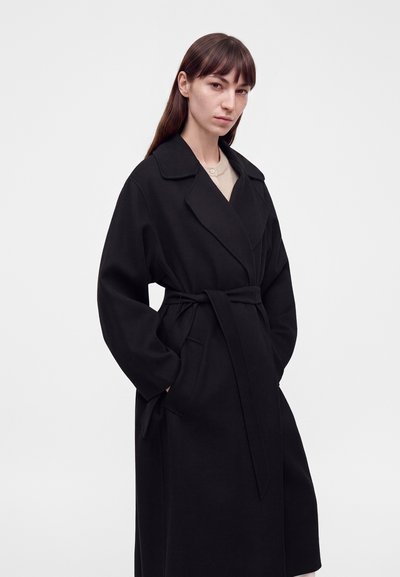 Femme aux longs cheveux bruns portant un manteau noir ceinturé par-dessus un haut clair, debout les mains dans les poches devant un fond blanc uni.