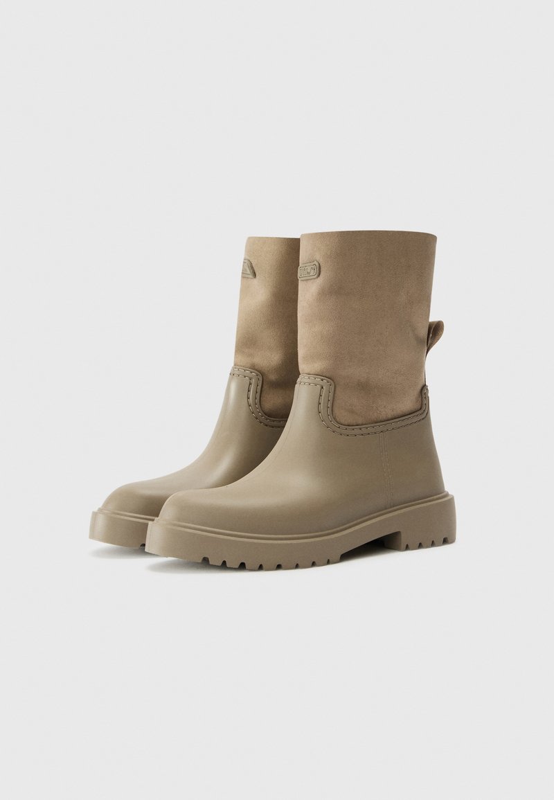 Botas beige hasta la mitad de la pantorrilla con parte inferior de goma, parte superior de ante, asa en el talón y suela gruesa, presentadas sobre un fondo blanco.