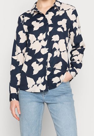 Femme portant une chemise boutonnée bleu foncé avec un grand motif de feuilles blanches et un jean bleu clair, une main dans la poche.