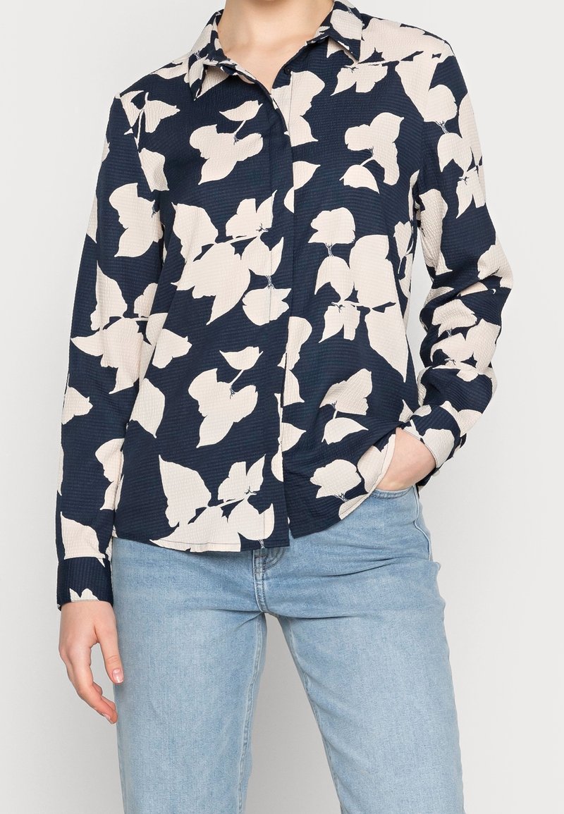 Femme portant une chemise boutonnée bleu foncé avec un grand motif de feuilles blanches et un jean bleu clair, une main dans la poche.