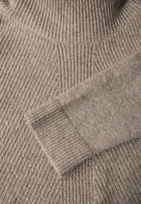 Beige stickad tröja med hög ribbad krage, som har texturerade mönster och en slät mudd. Mjuk, varm tyg med detaljerad sömnad synlig.