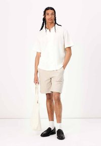 Hvid kortærmet skjorte med vertikal struktur, beige stribede shorts, sorte loafers, hvide sokker og en lysefarvet tote bag.